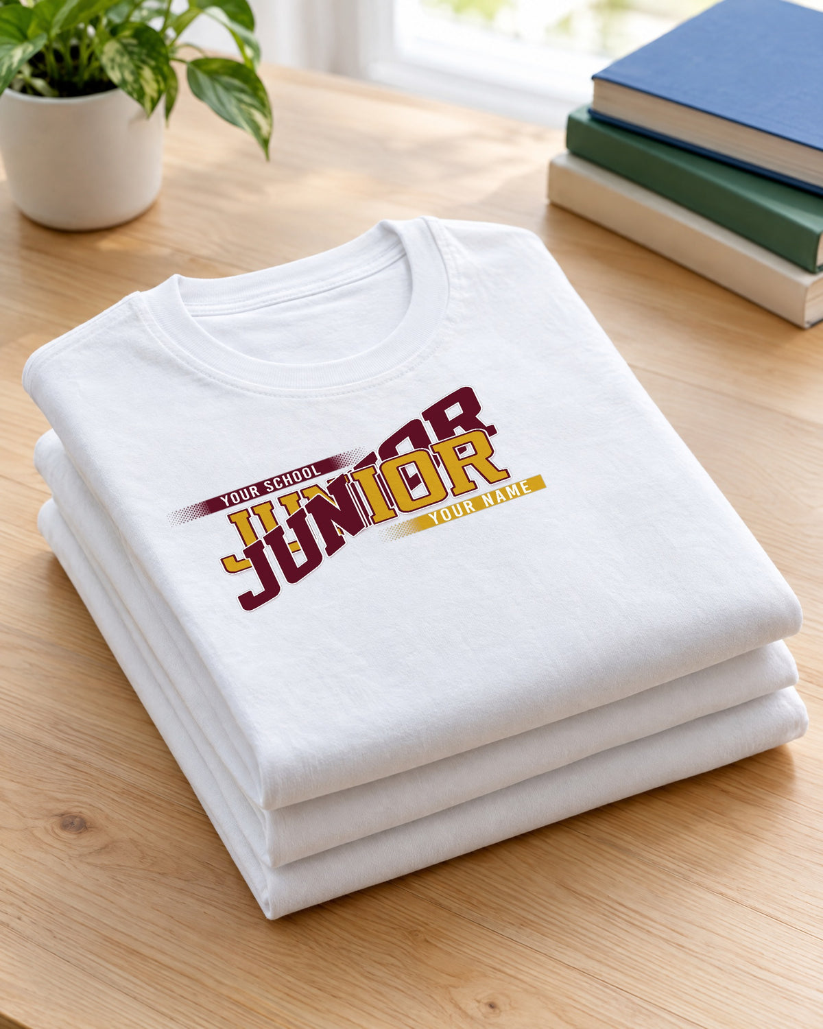 Folded stack of white custom Junior class year shirts in maroon and gold school colors on a wood table