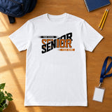 White custom Senior class year shirt in burnt orange and black laid flat on a wood desk with a school backpack, notebook, pencils and ID lanyard