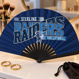 Navy game day clack fan with black ribs and a volleyball team design on a white vanity with makeup brushes and gold hoop earrings.