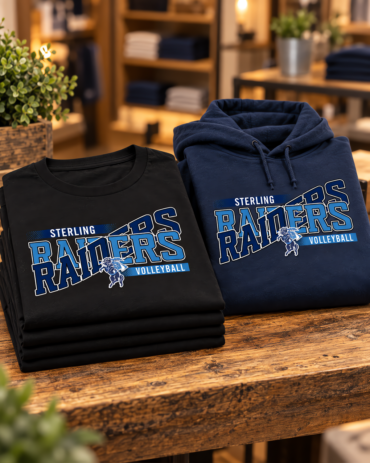 Folded stack of black game day tees and a navy game day hoodie with a volleyball team design on a wooden store table.