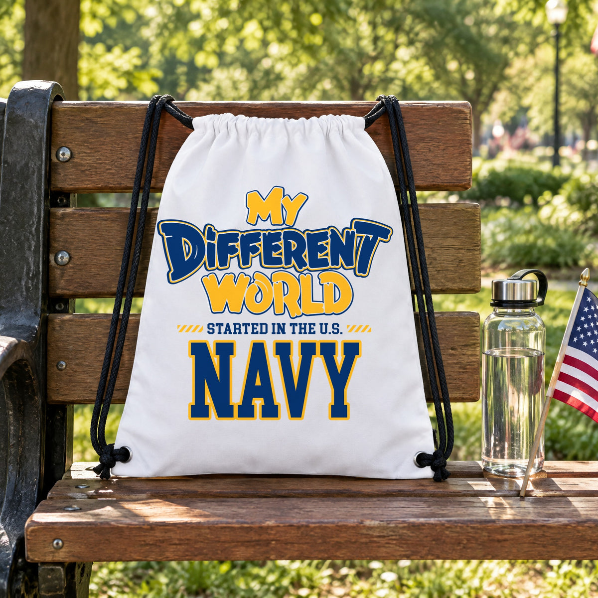 My Different World Started in the U.S. Navy white drawstring backpack hanging on a wooden park bench, with a water bottle and a small American flag