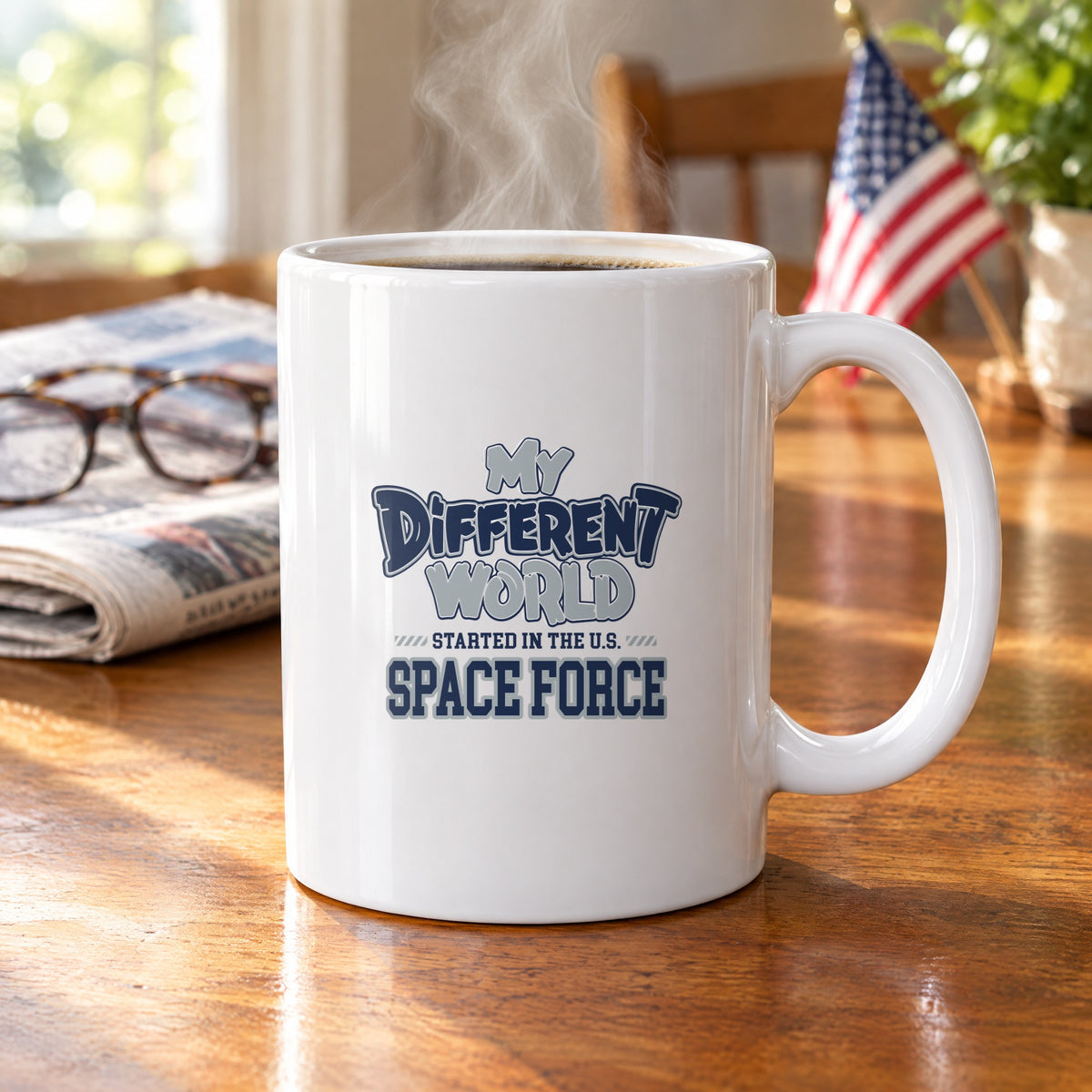 My Different World Started in the U.S. Space Force coffee mug with hot coffee on a wooden kitchen table, with reading glasses, a newspaper and a small American flag